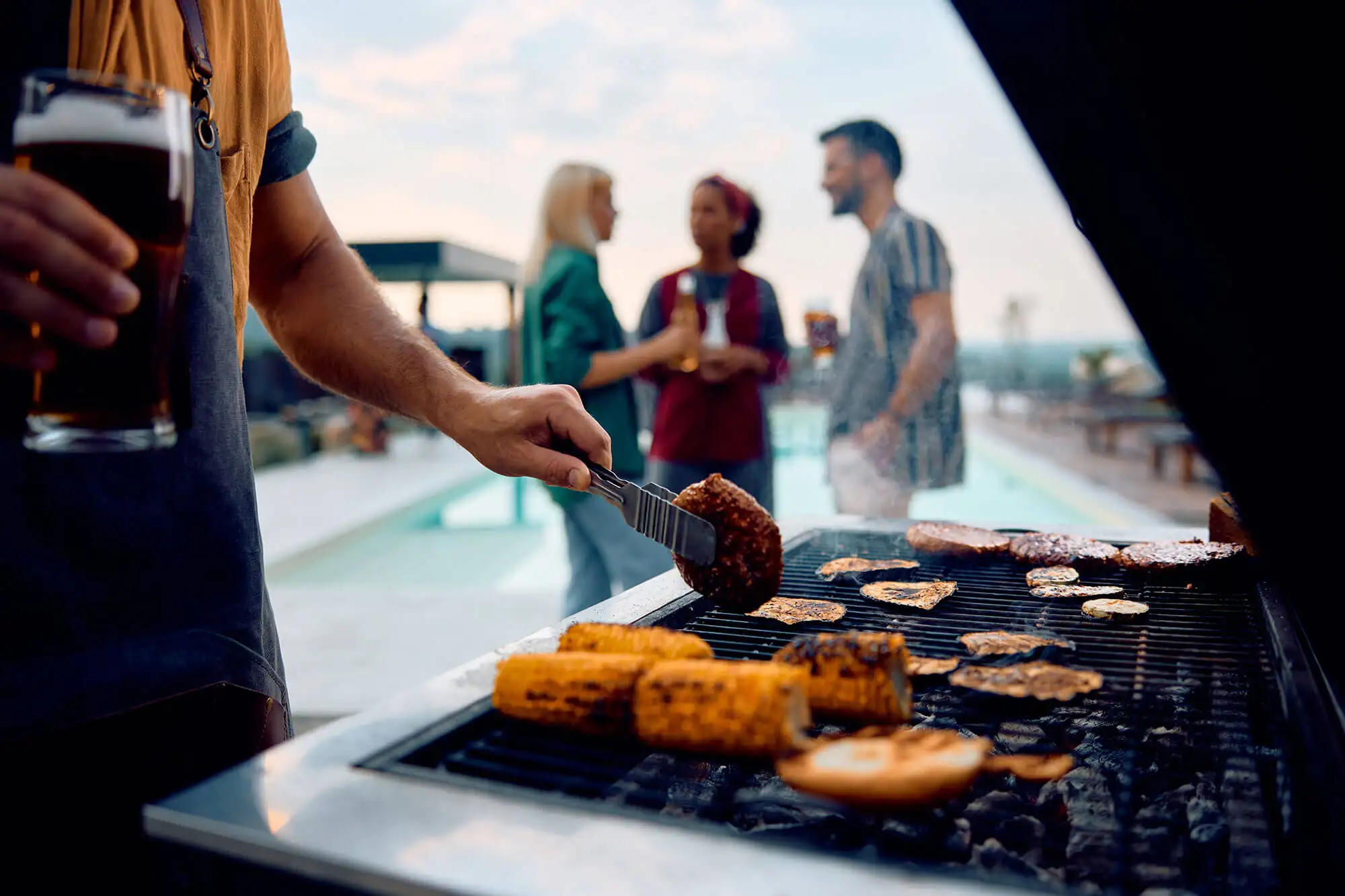 Man holding a beer and grilling burgers with friends in the background by the pool